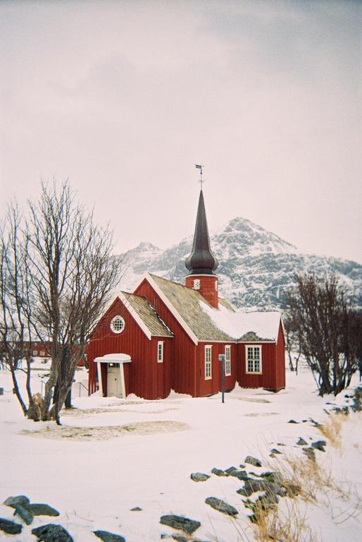 A red church with a steeple stands in a snowy landscape with mountains in the background.