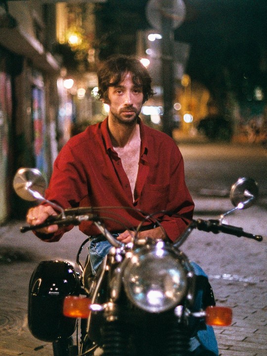 A man in a red shirt sits on a motorcycle at night on a street.
