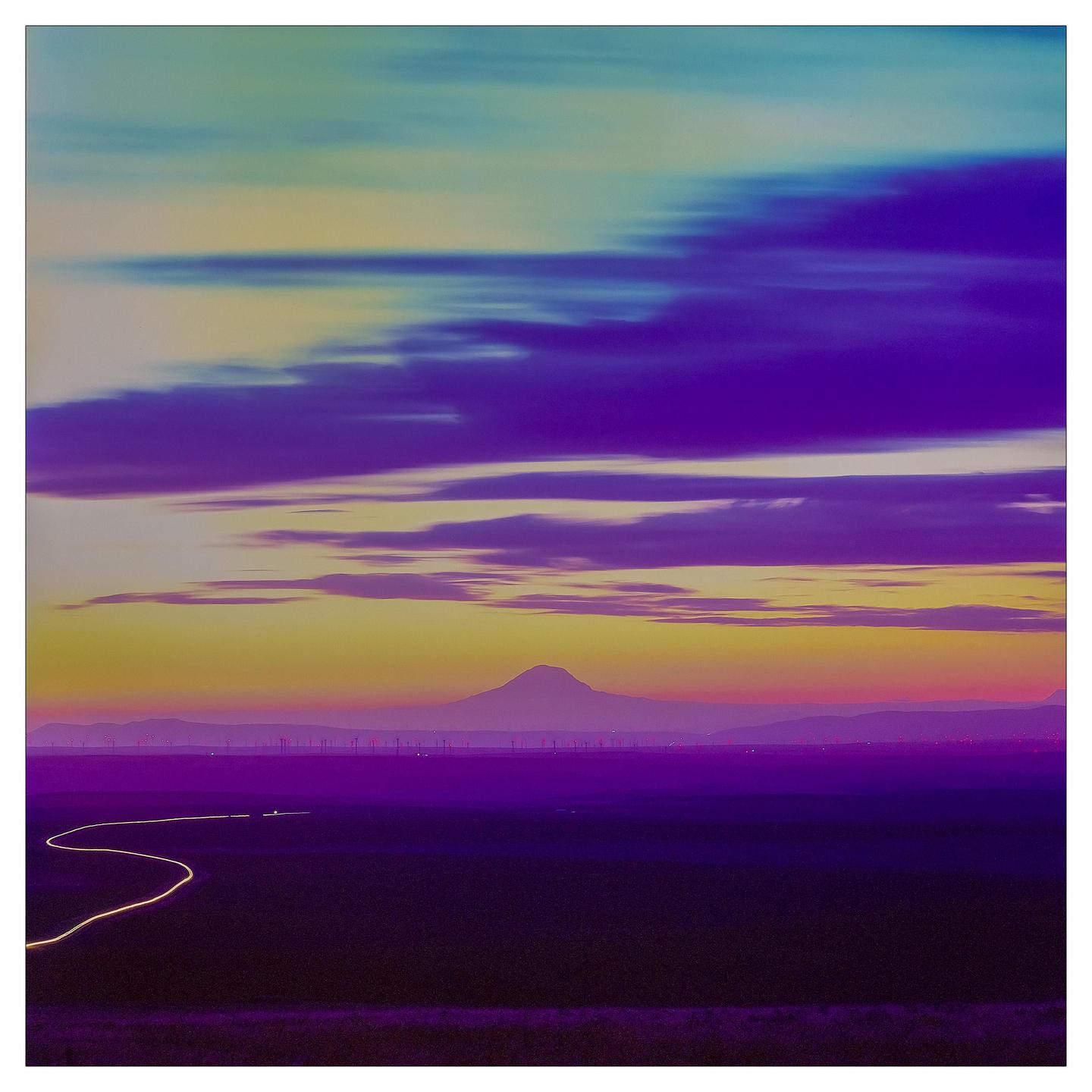 A winding light trail crosses a field towards a mountain range under a vibrant, cloudy sky.
