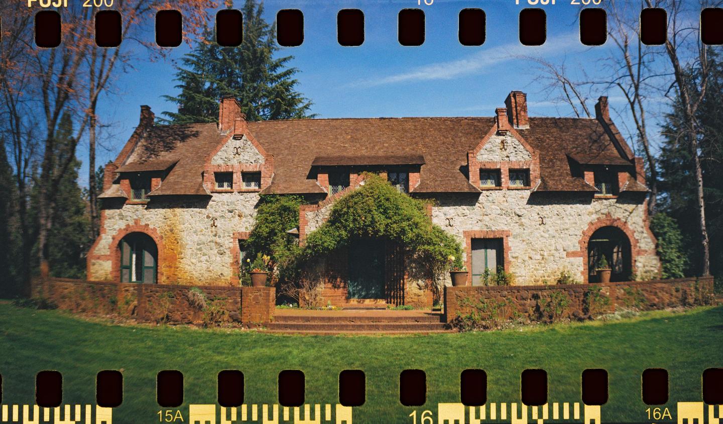 A stone and brick house with a brown roof and green ivy growing on its facade is surrounded by a green lawn and trees under a clear blue sky.