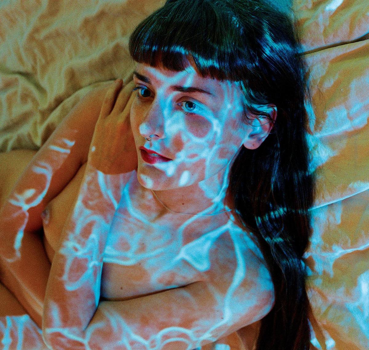 A woman with long dark hair and bangs lies on a bed with blue light patterns projected onto her body.