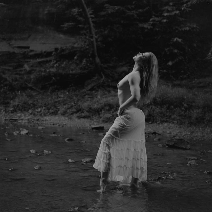 A topless woman in a long skirt stands in a shallow stream, looking up with her hair flowing back.