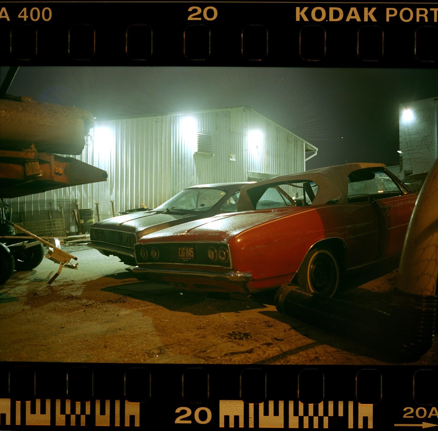 Two vintage cars are parked at night next to a corrugated metal building with bright lights.