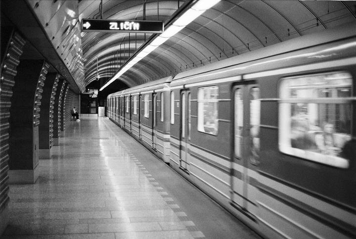 A subway train speeds through a station with a man sitting on a bench in the distance.
