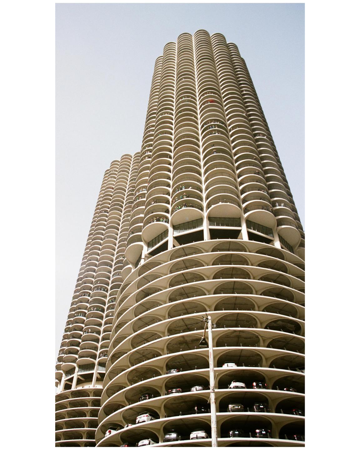 Two tall, cylindrical buildings with many balconies and visible parking levels are shown from a low angle against a light sky.