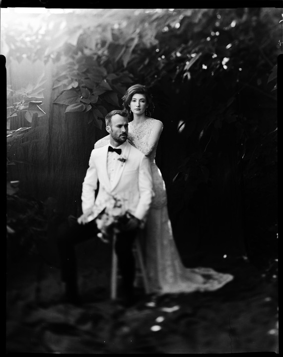 A man in a tuxedo sits while a woman in a wedding dress stands behind him, both looking in different directions.
