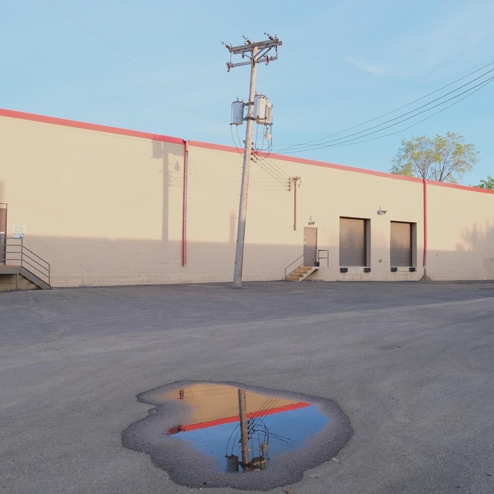 A utility pole stands in front of a building with a red stripe, reflected in a puddle on the asphalt.