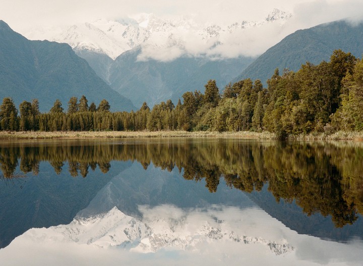 A lake reflects a forest and snow-capped mountains under a cloudy sky.