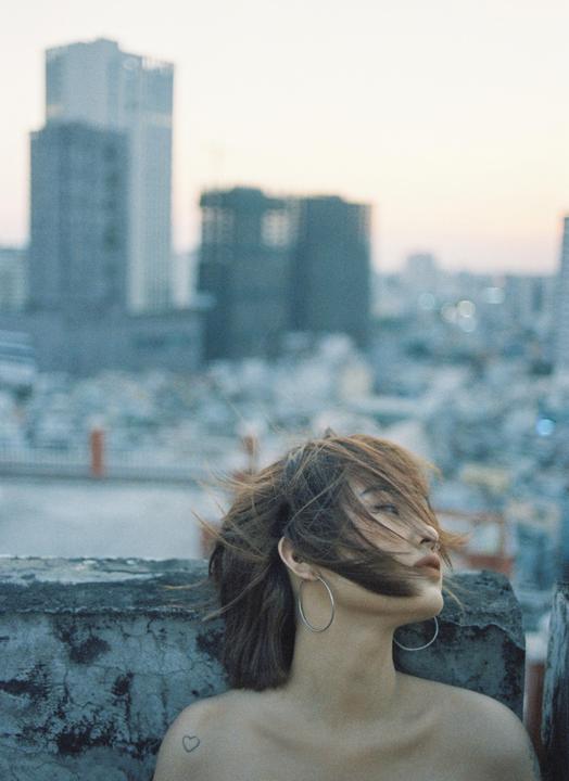 A woman with windblown hair and hoop earrings looks out over a city skyline at dusk.