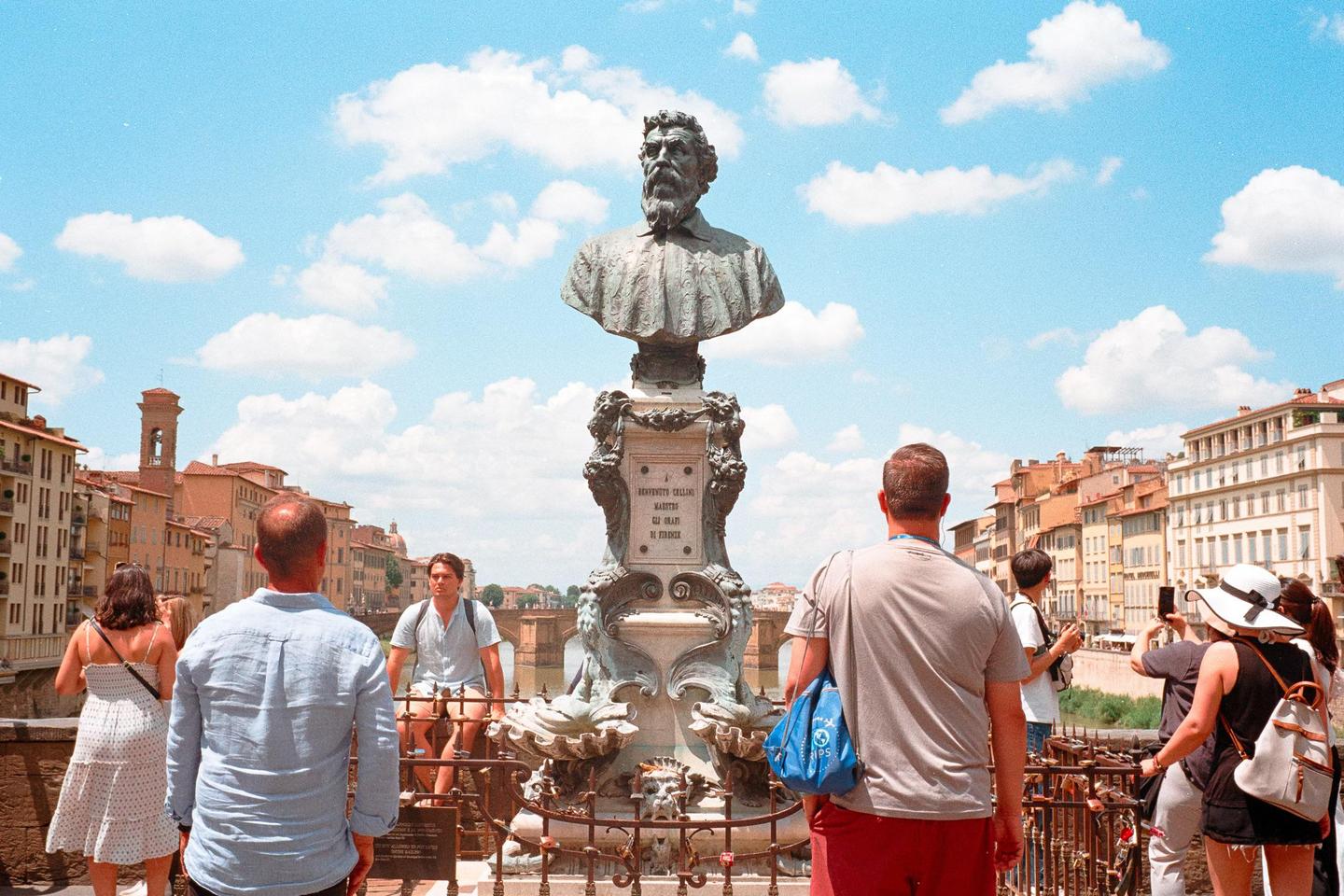 A bust of a man on a pedestal stands on a bridge with people looking at it and the buildings in the background.
