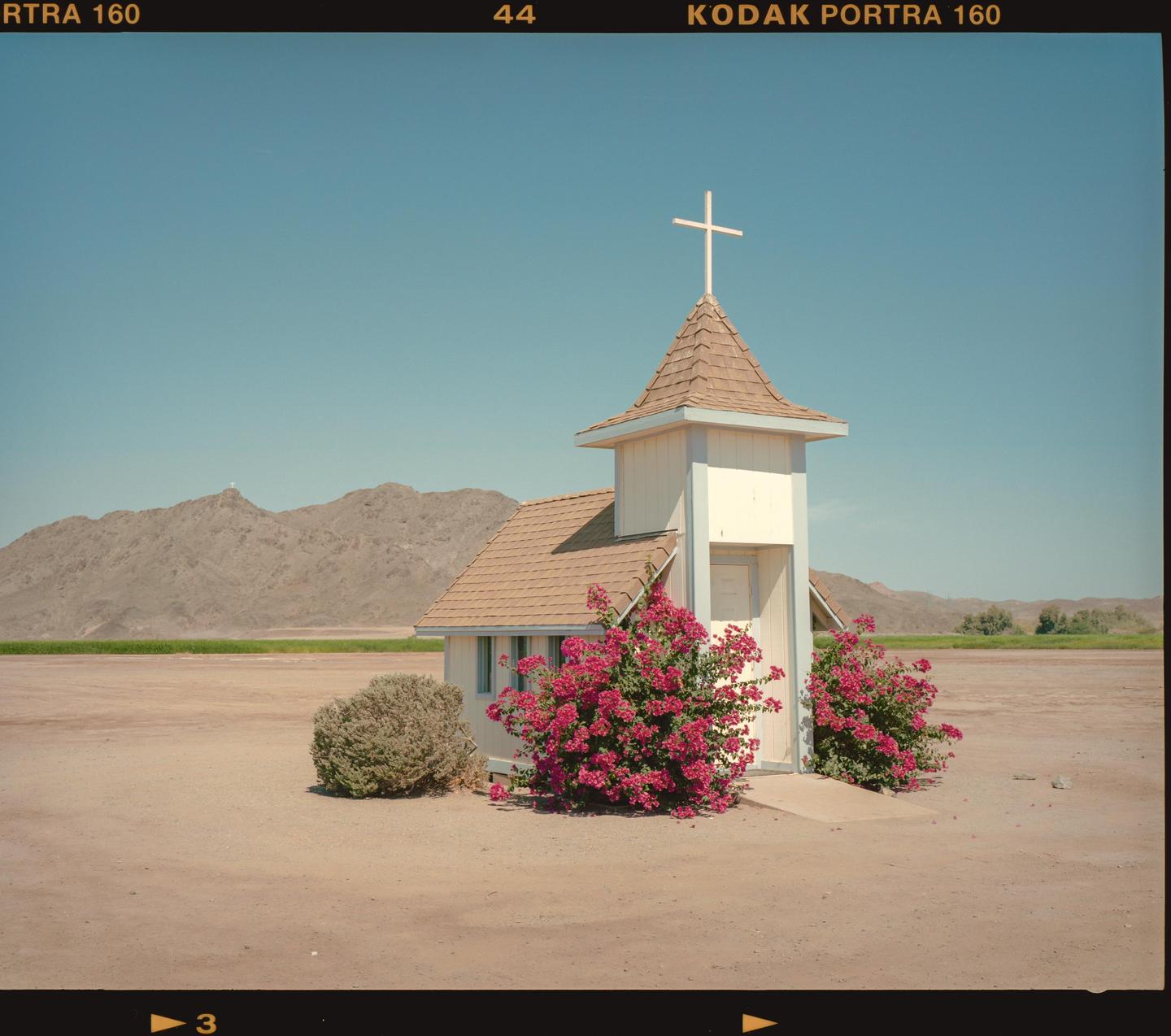 A small chapel with a cross on its steeple stands in a desert landscape with mountains in the background.