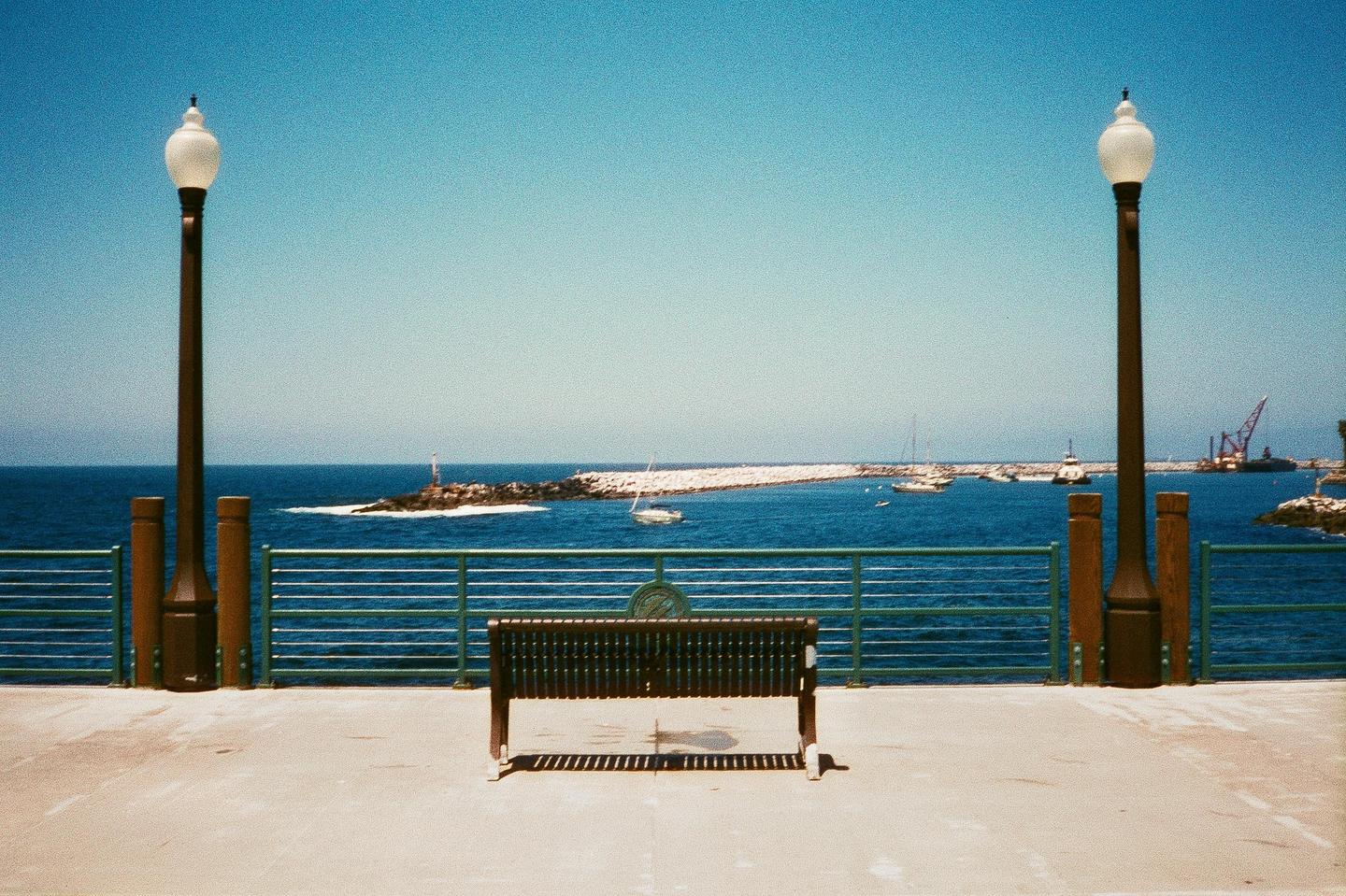 A lone bench sits on a pier overlooking a harbor with boats and a breakwater under a clear sky.