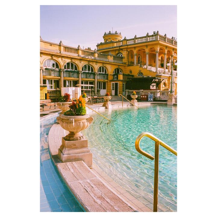 A large yellow building with arched windows overlooks a swimming pool with clear blue water and a golden handrail.
