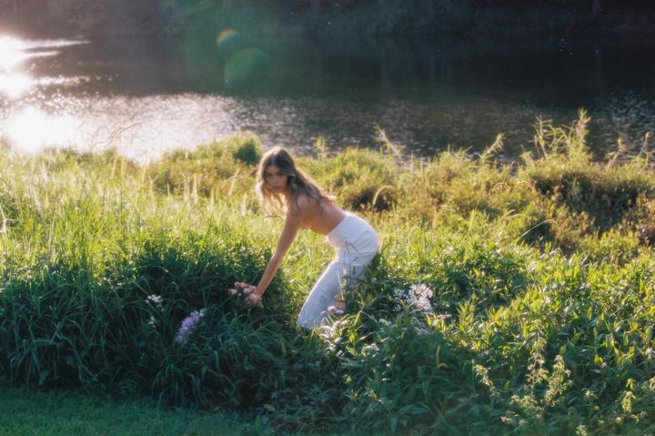 A woman in white pants is picking flowers in a field next to a body of water.