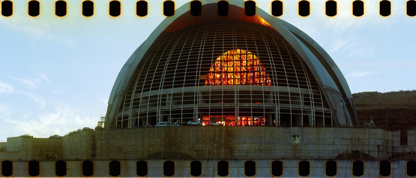 A large domed building with a grid pattern and a section of orange stained glass is visible under a clear sky.