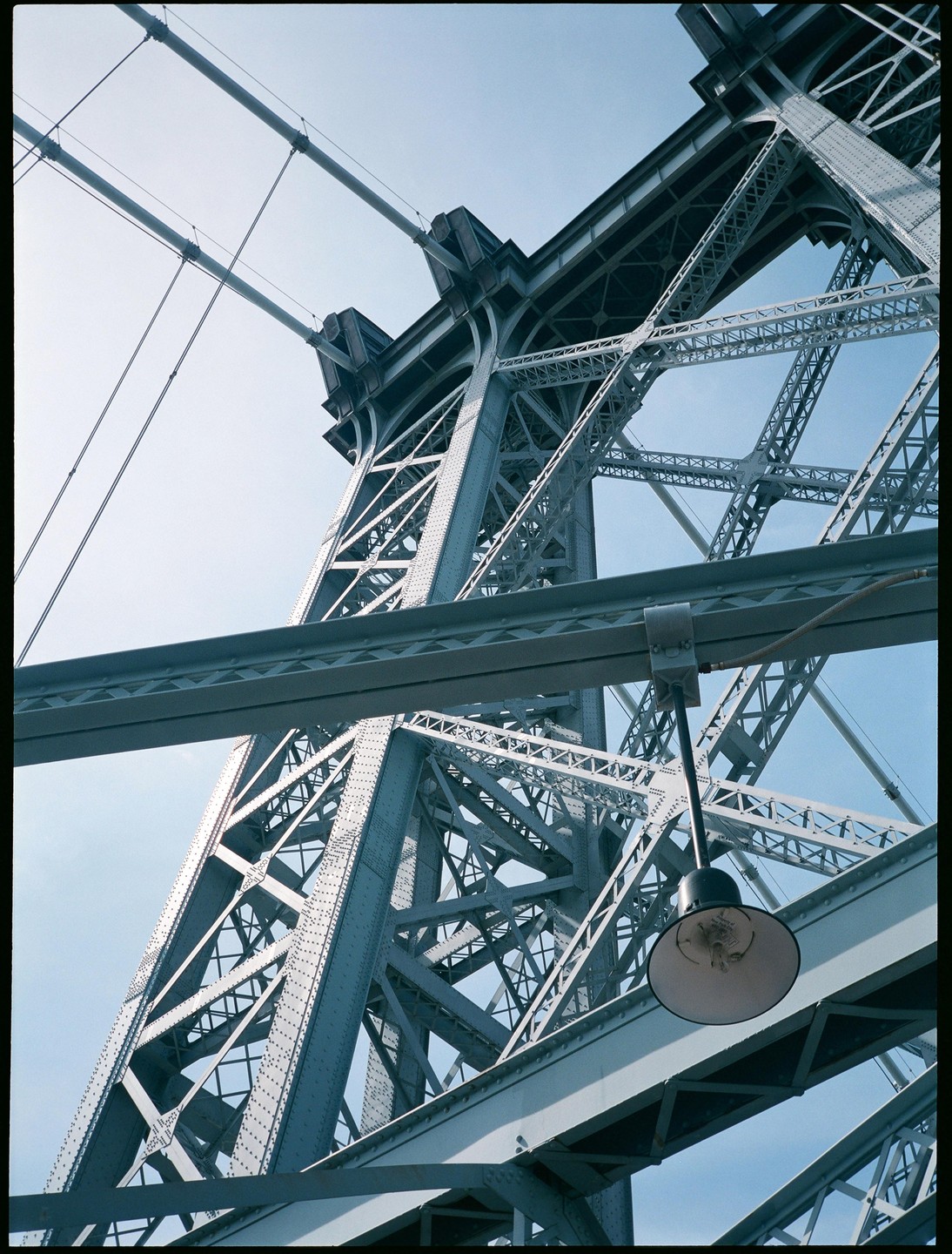 A low-angle view shows the intricate metal structure of a bridge against a clear sky, with a single lamp hanging in the foreground.