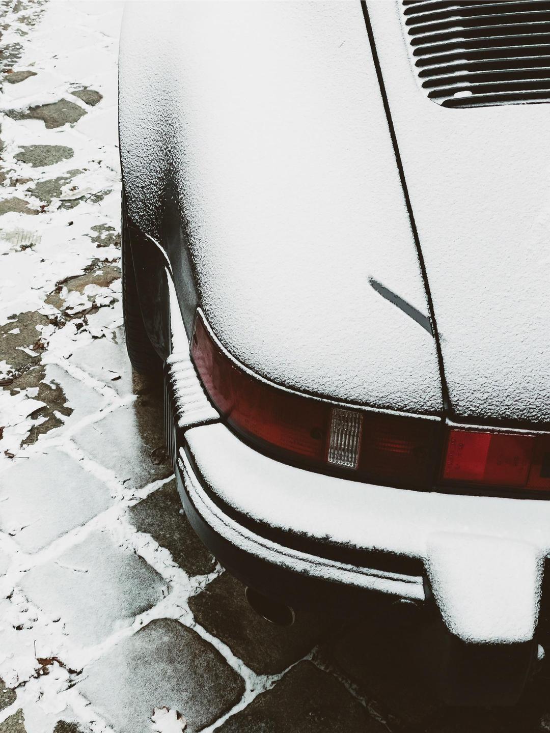 The rear of a car is covered in snow and parked on a cobblestone street.