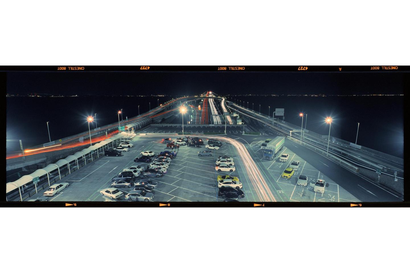 A long exposure captures light trails from vehicles on a bridge and in a parking area at night.