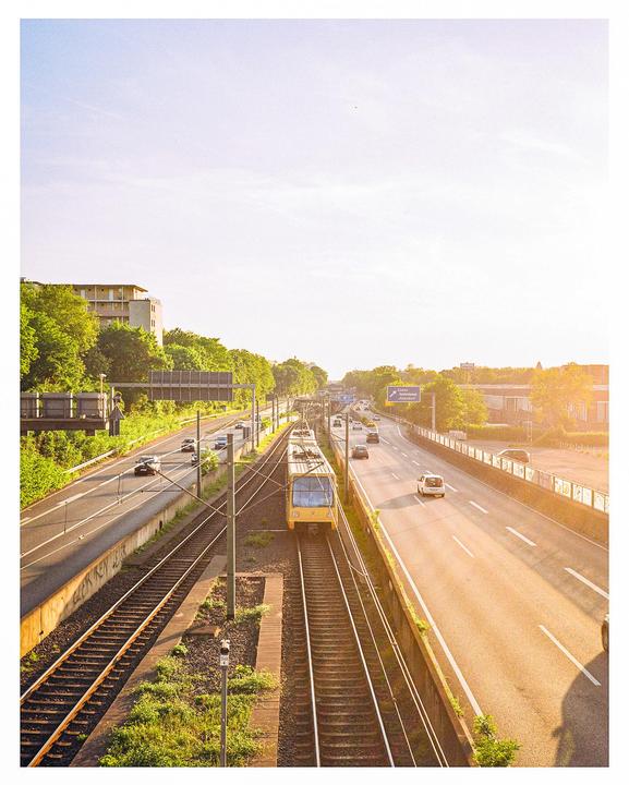 A yellow train travels on tracks parallel to a highway with cars under a bright sky.