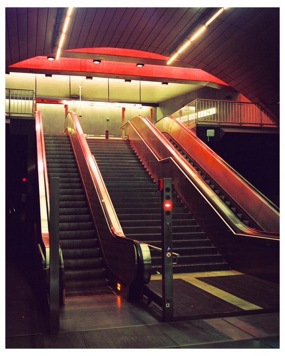 An escalator and a staircase lead up to a subway platform with red lighting.