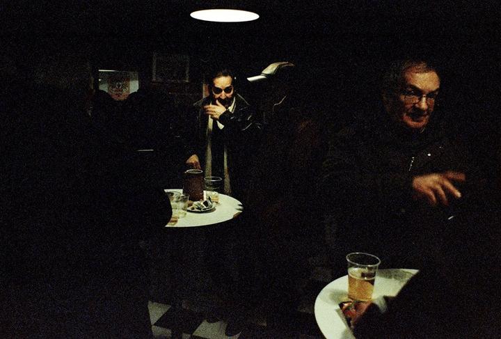 A man in sunglasses stands near a table with drinks in a dimly lit room while another man sits at a separate table with a drink.
