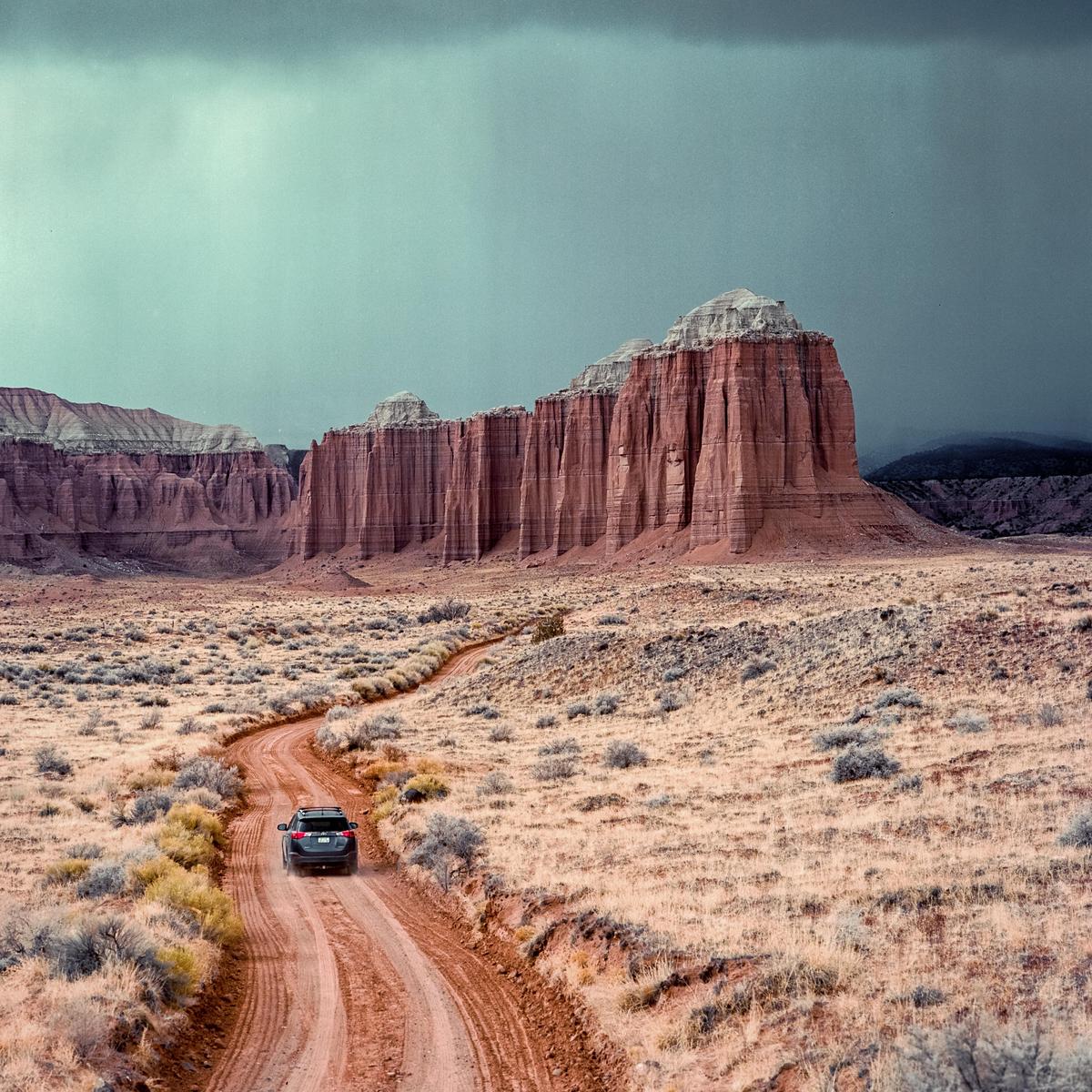 A car drives on a dirt road through a desert landscape with large rock formations under a cloudy sky.