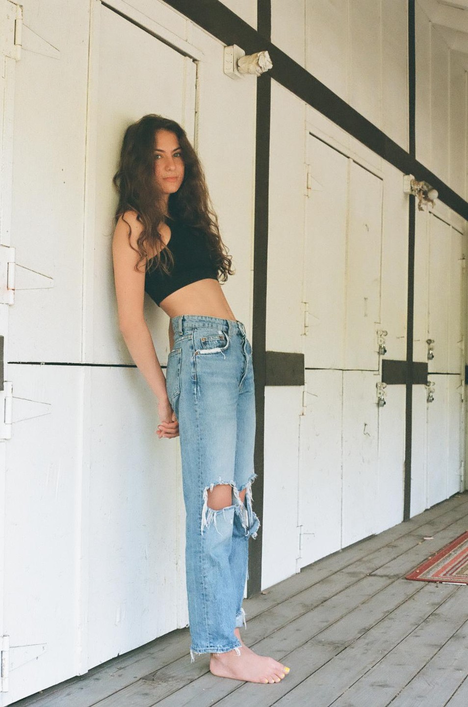 A young woman with long, curly hair leans against a white wall with black trim, wearing a black crop top and ripped blue jeans.