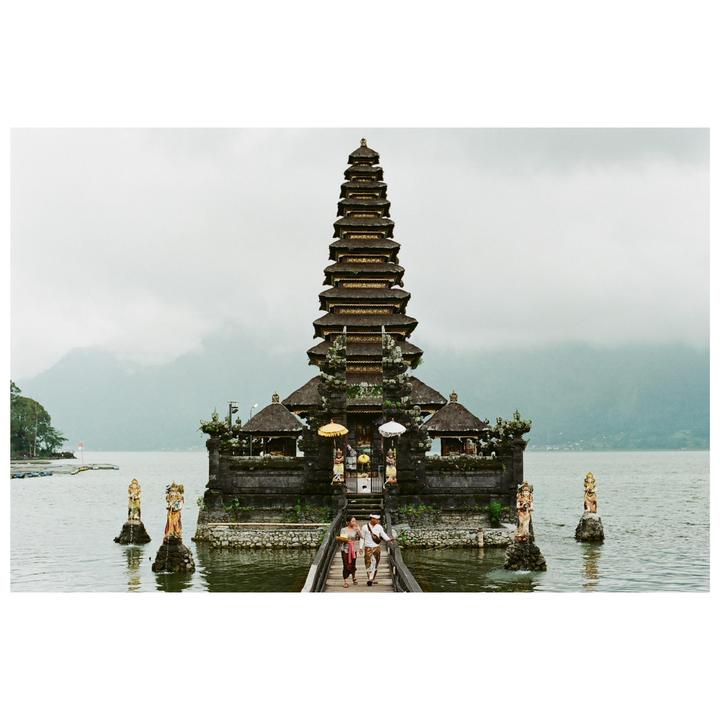 A couple walks on a wooden bridge towards a multi-tiered temple situated on a lake with mountains in the background.