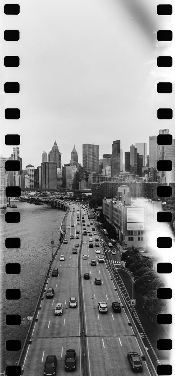 A black and white high-angle shot shows a multi-lane highway with cars next to a body of water, with a city skyline in the background.