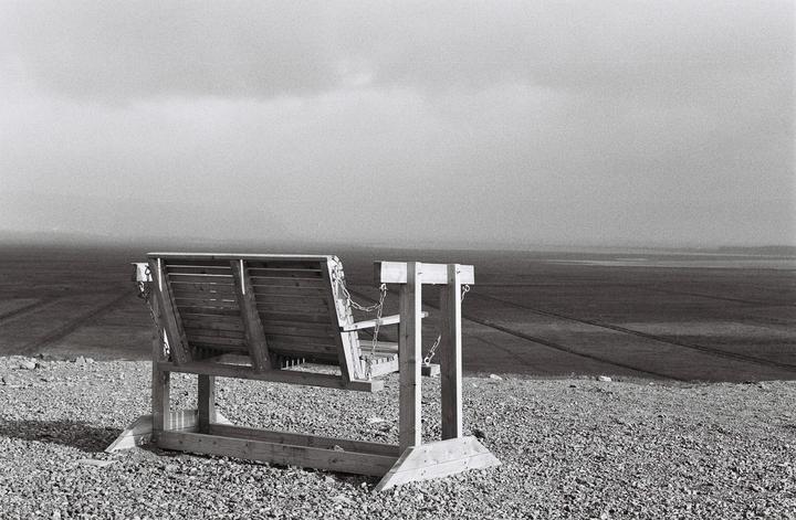 A wooden swing bench sits on a gravelly hill overlooking a vast, flat landscape under a cloudy sky.