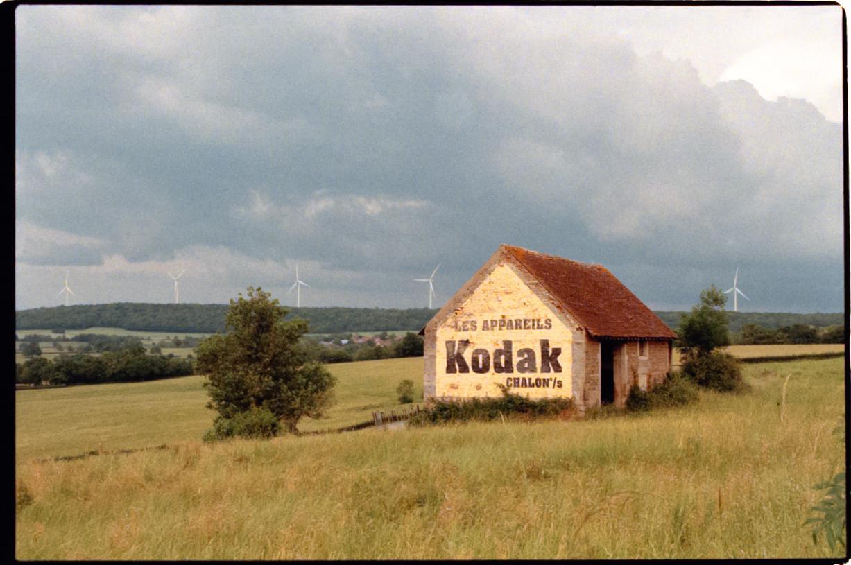 An old building with a Kodak advertisement painted on its side stands in a grassy field under a cloudy sky, with wind turbines visible in the distance.