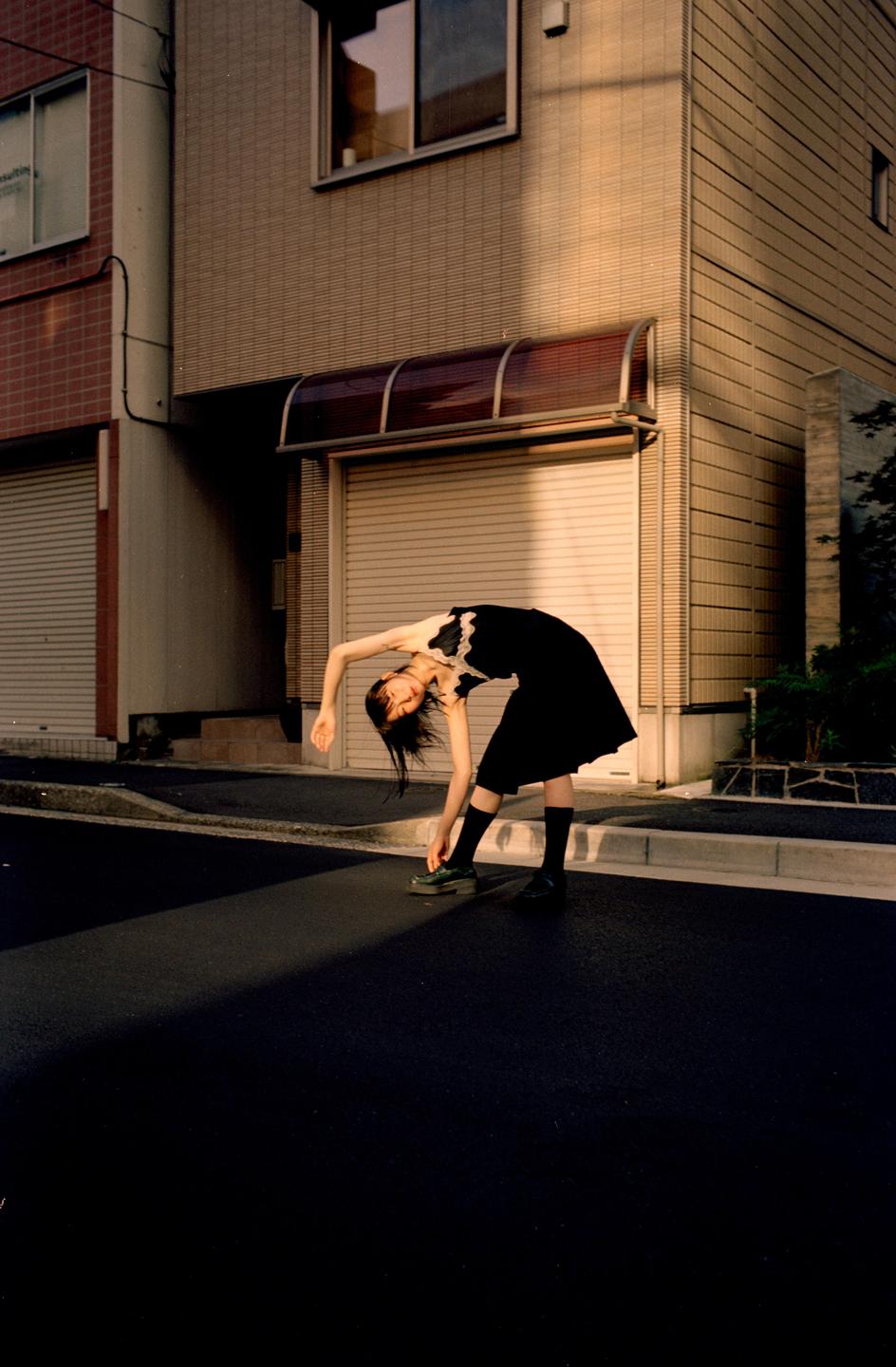A woman in a black dress bends over to tie her shoe on a street with buildings in the background.
