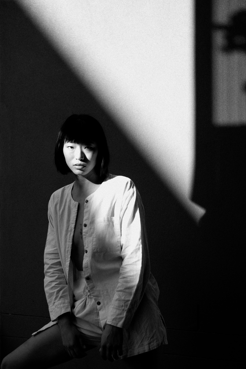 A woman with short dark hair and an unbuttoned shirt sits in a room with strong shadows.