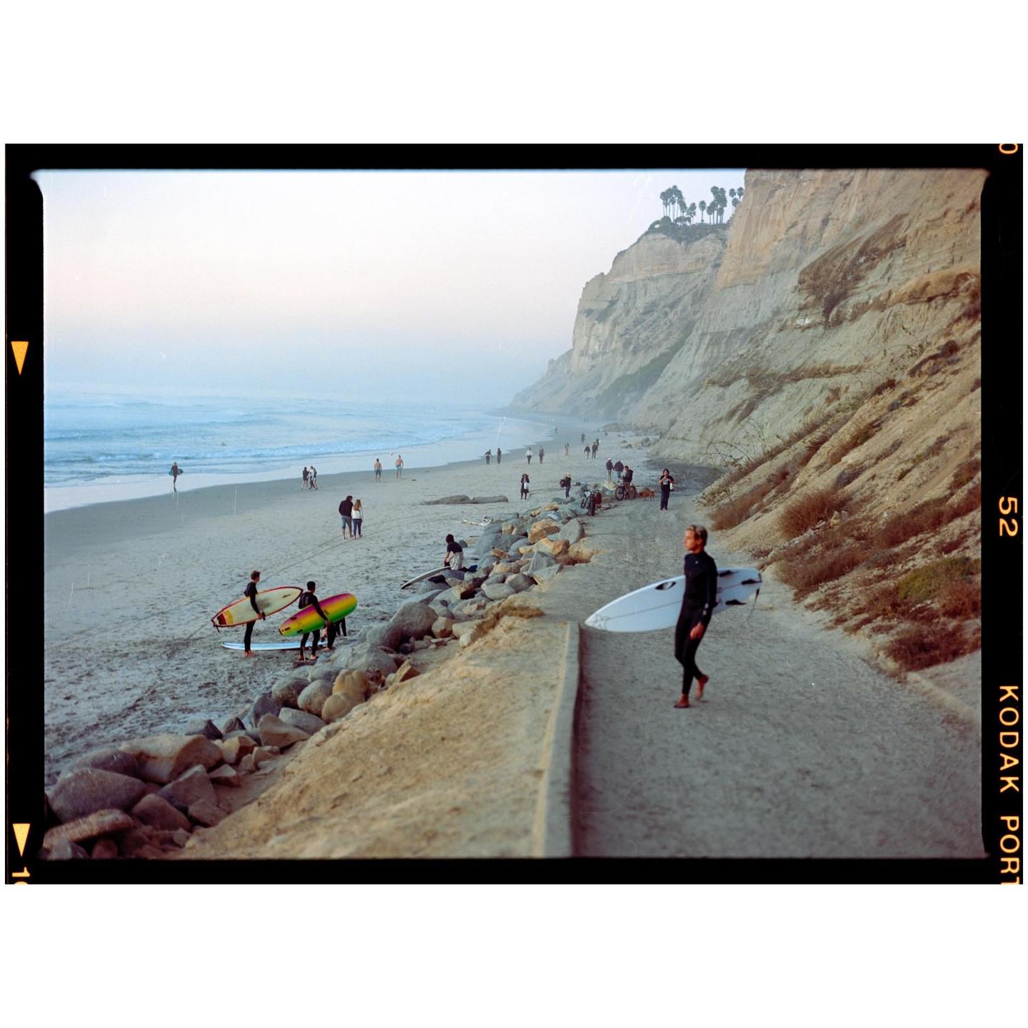 A surfer walks along a path next to a beach with cliffs in the background.