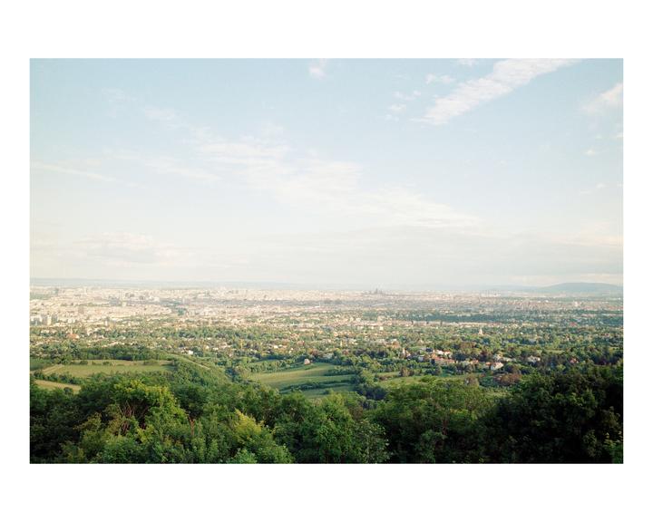 A panoramic view of a city with many buildings and green trees is seen from a high vantage point under a cloudy sky.