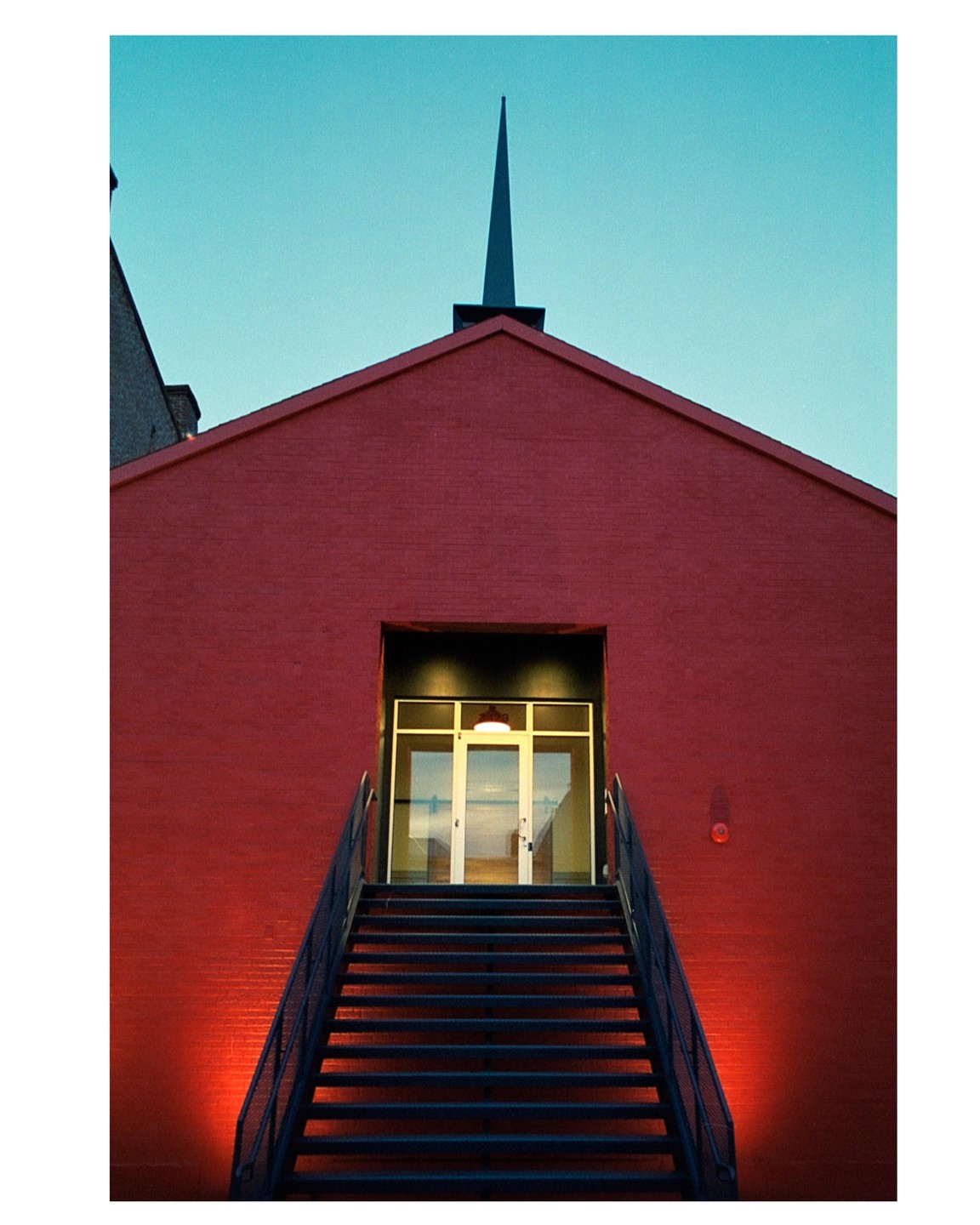 A red brick building with a steeple and a set of stairs leading to an illuminated entrance is shown under a teal sky.