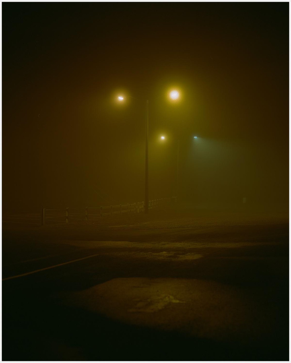 Three streetlights illuminate a foggy night scene with a fence and puddles on the ground.
