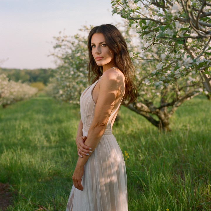 A woman in a light dress stands in a blooming orchard with green grass and trees with white flowers.