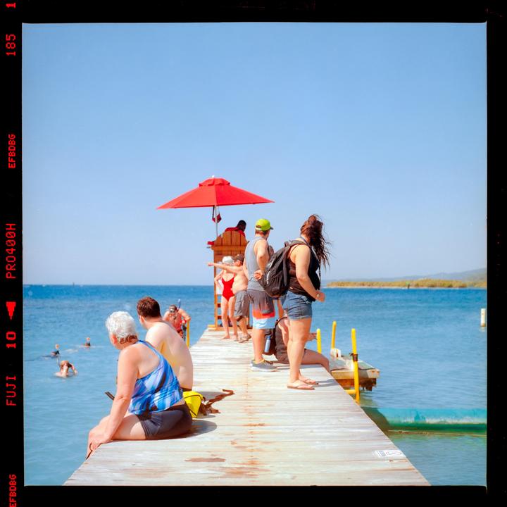 A group of people are on a wooden pier next to a body of water with a red umbrella and lifeguard stand.