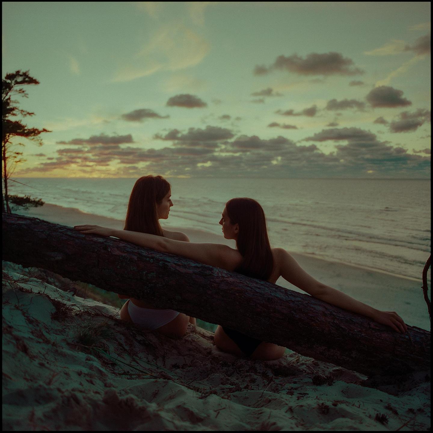 Two women sit on a sandy beach next to a fallen tree trunk, looking at each other with the ocean and a cloudy sky in the background at sunset.