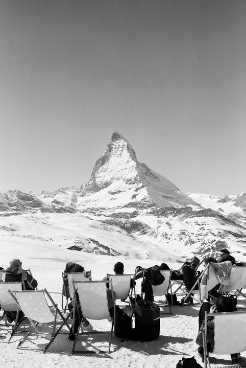 People sit in chairs in the snow, looking at a large snow-covered mountain.