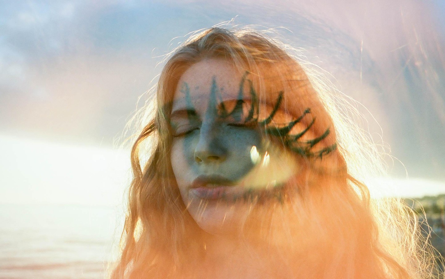 A woman with closed eyes and painted face is shown in a double exposure with a bright sky and water.