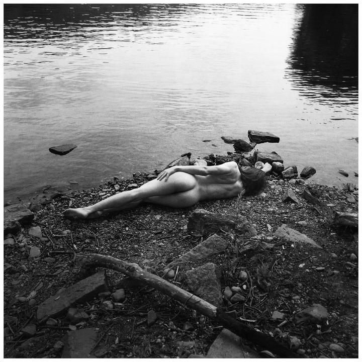 A nude woman lies on a rocky shore next to a body of water.
