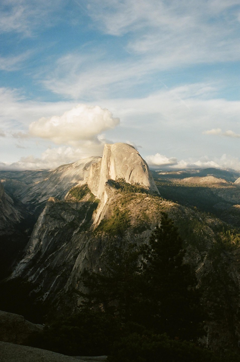 A large granite dome rises above a valley with forested mountains under a partly cloudy sky.
