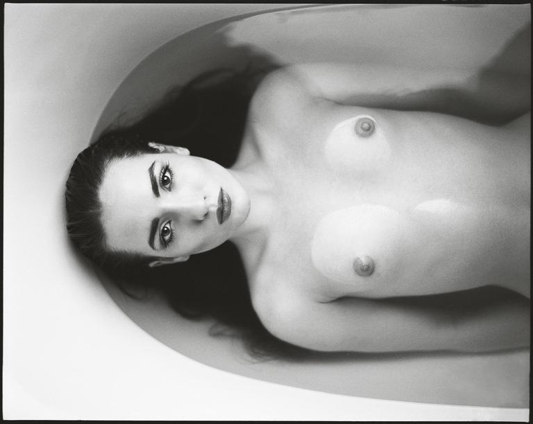 A woman with long hair is lying in a bathtub, looking up at the viewer.