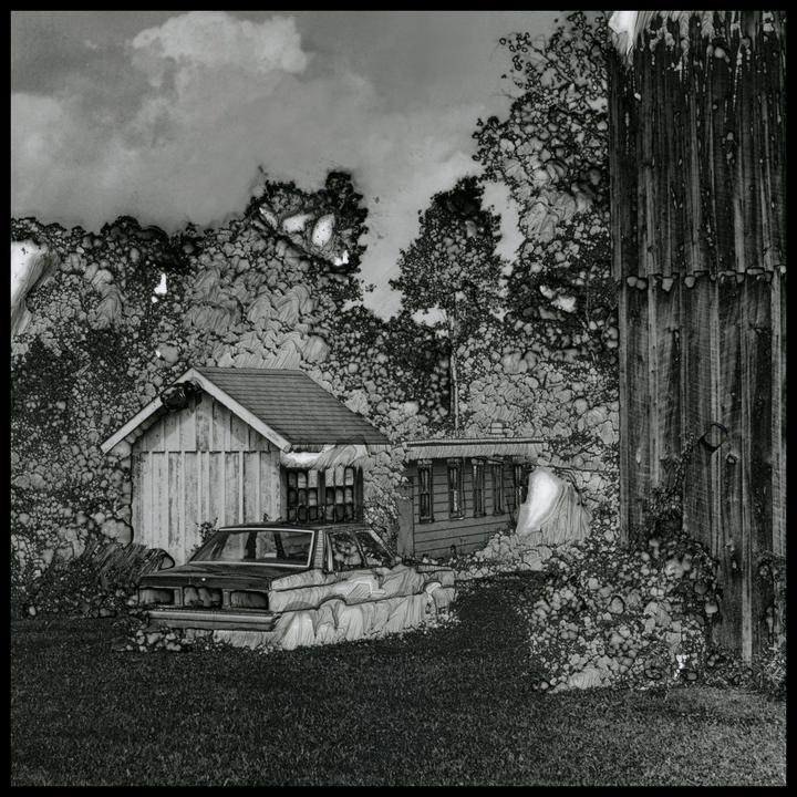 An old car is parked in front of two small buildings with a tall wooden structure on the right.