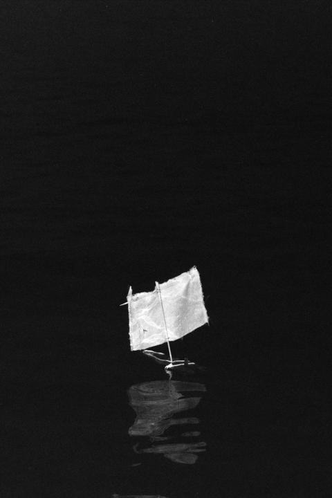 A small toy sailboat with a white sail floats on dark water, reflecting on the surface.