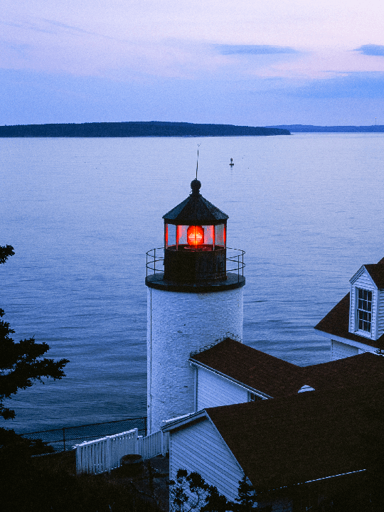 A lighthouse with a glowing red light stands by the ocean at dusk.