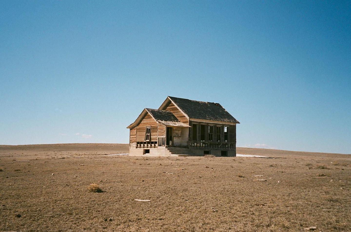 An abandoned wooden house stands alone in a vast, dry field under a clear blue sky.