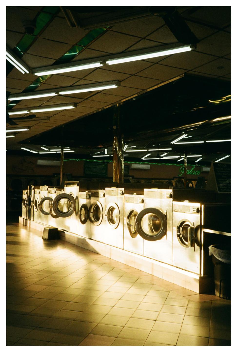 A row of washing machines with open doors lines a laundromat with fluorescent lighting and tiled floors.
